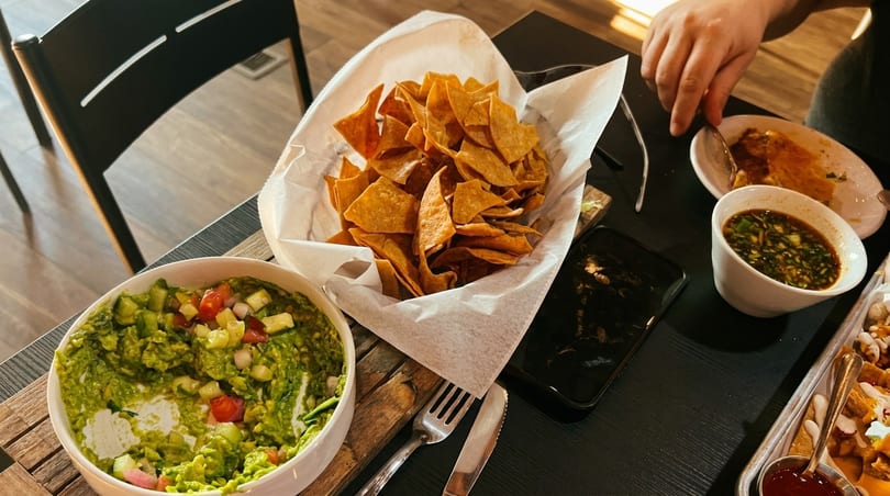 A table with a large bowl of guacamole, a basket of tortilla chips, and a person eating.