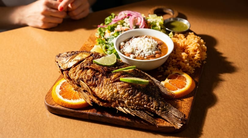 Crispy fried whole fish with lime, rice, beans, salad, and sauce on a wooden board.