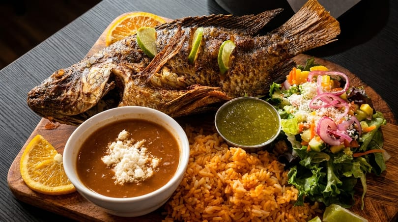 Whole fried fish with lime slices, rice, beans, salsa, and salad on a wooden board.