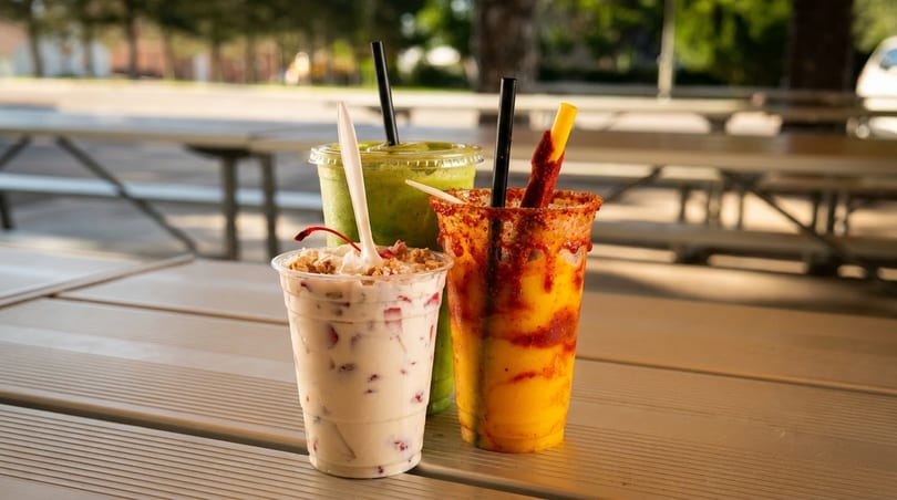 Three colorful drinks on a picnic table: a strawberry shake, a green smoothie, and a spicy mango drink.